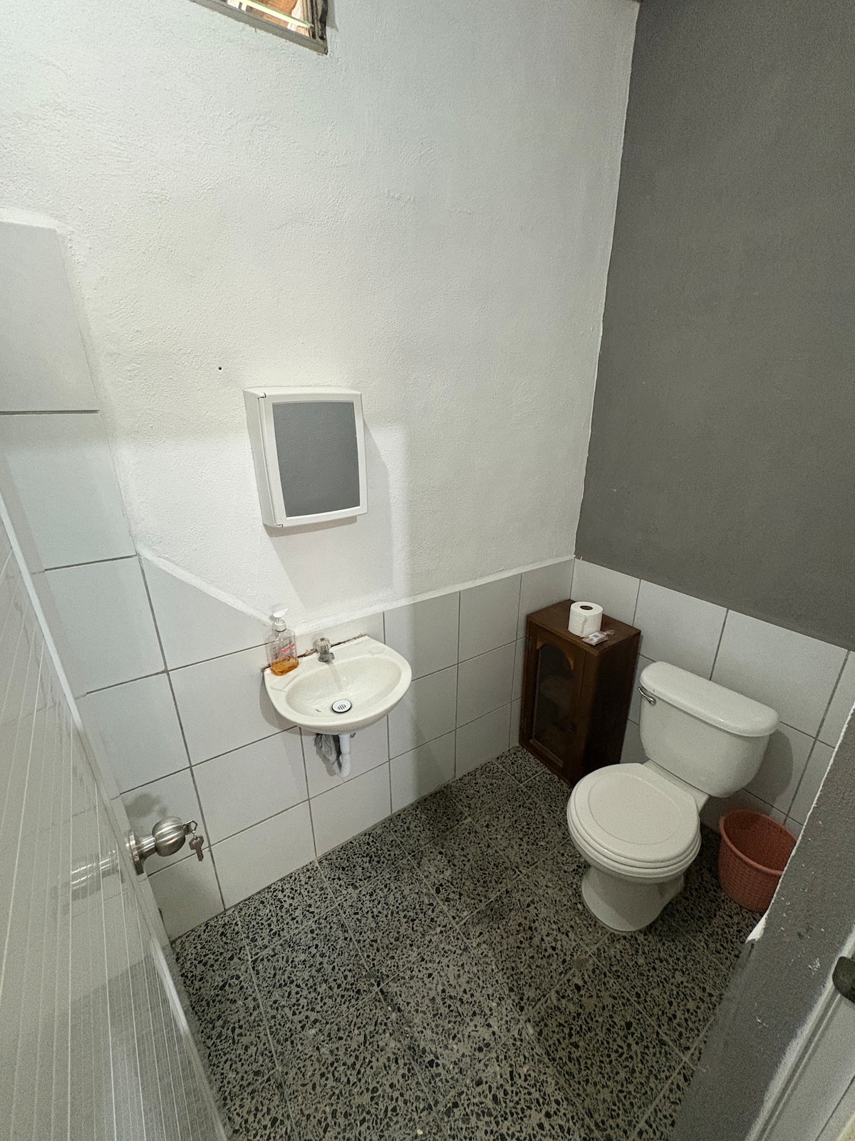 A compact bathroom features white tiled walls and a mosaic floor. A small sink is positioned opposite a toilet, with a mirror mounted above the sink. A towel holder and toilet paper roll are visible, alongside a decorative wooden cabinet.