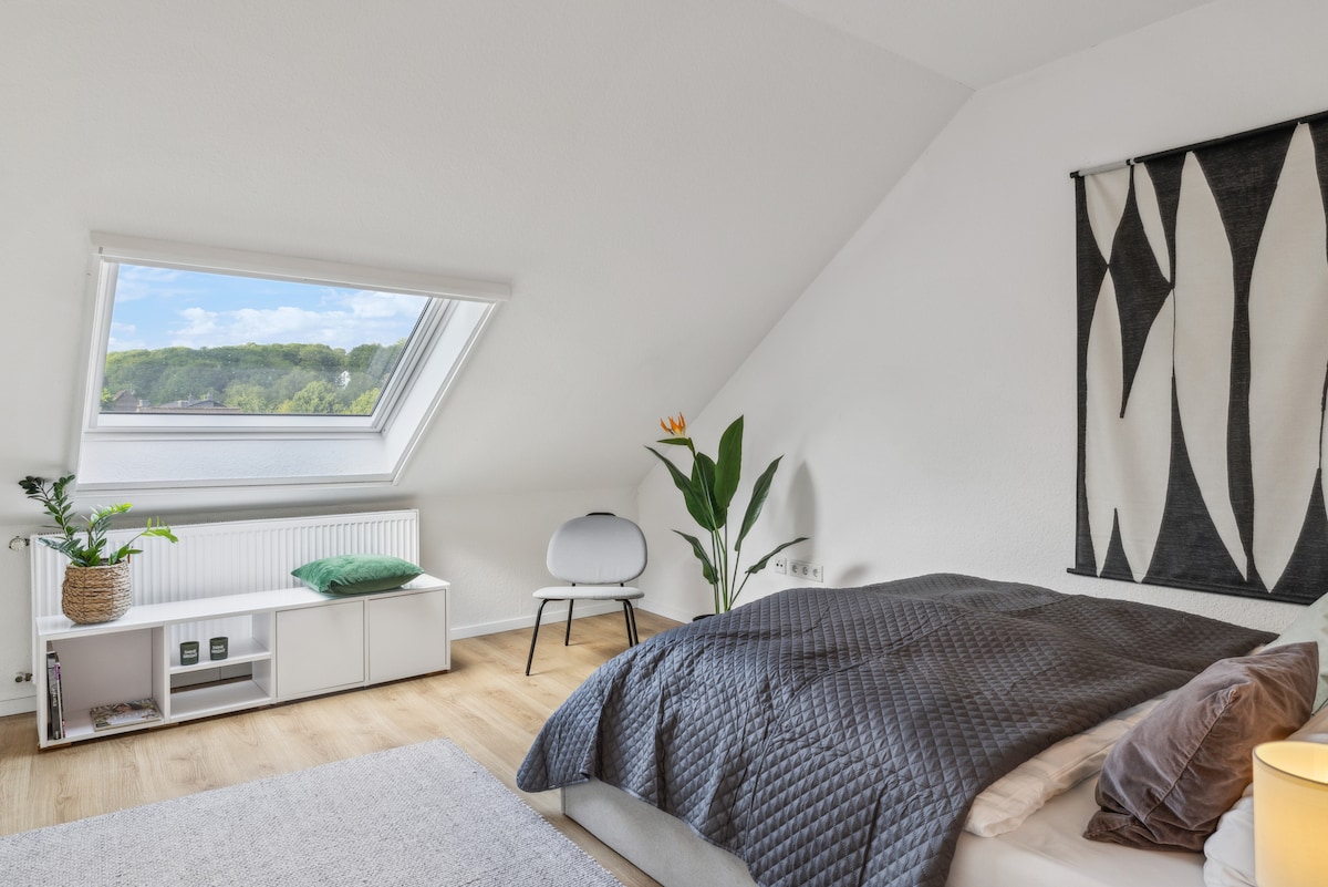A bright and airy bedroom features a sloped ceiling with a skylight, providing natural light and scenic views. A spacious bed is layered with a textured duvet, while a potted plant adds a touch of greenery. A minimalist chair and side table complete the welcoming atmosphere.