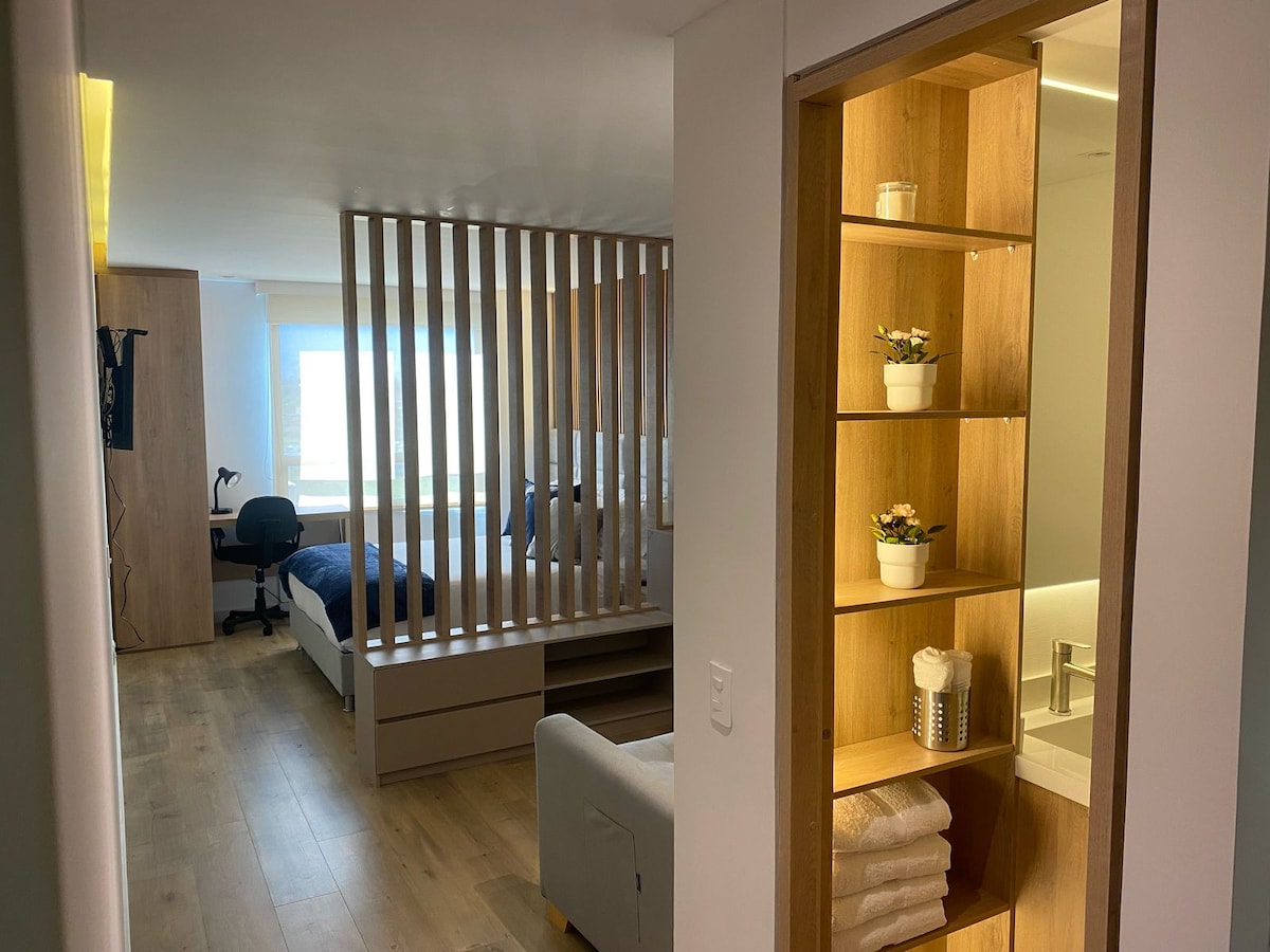 A hallway offers a view into a bright studio apartment. A spacious sleeping area features a bed behind a wooden slatted partition. An office chair is positioned next to a window. A small shelf displays potted plants and neatly stacked towels, with a bathroom area visible nearby.