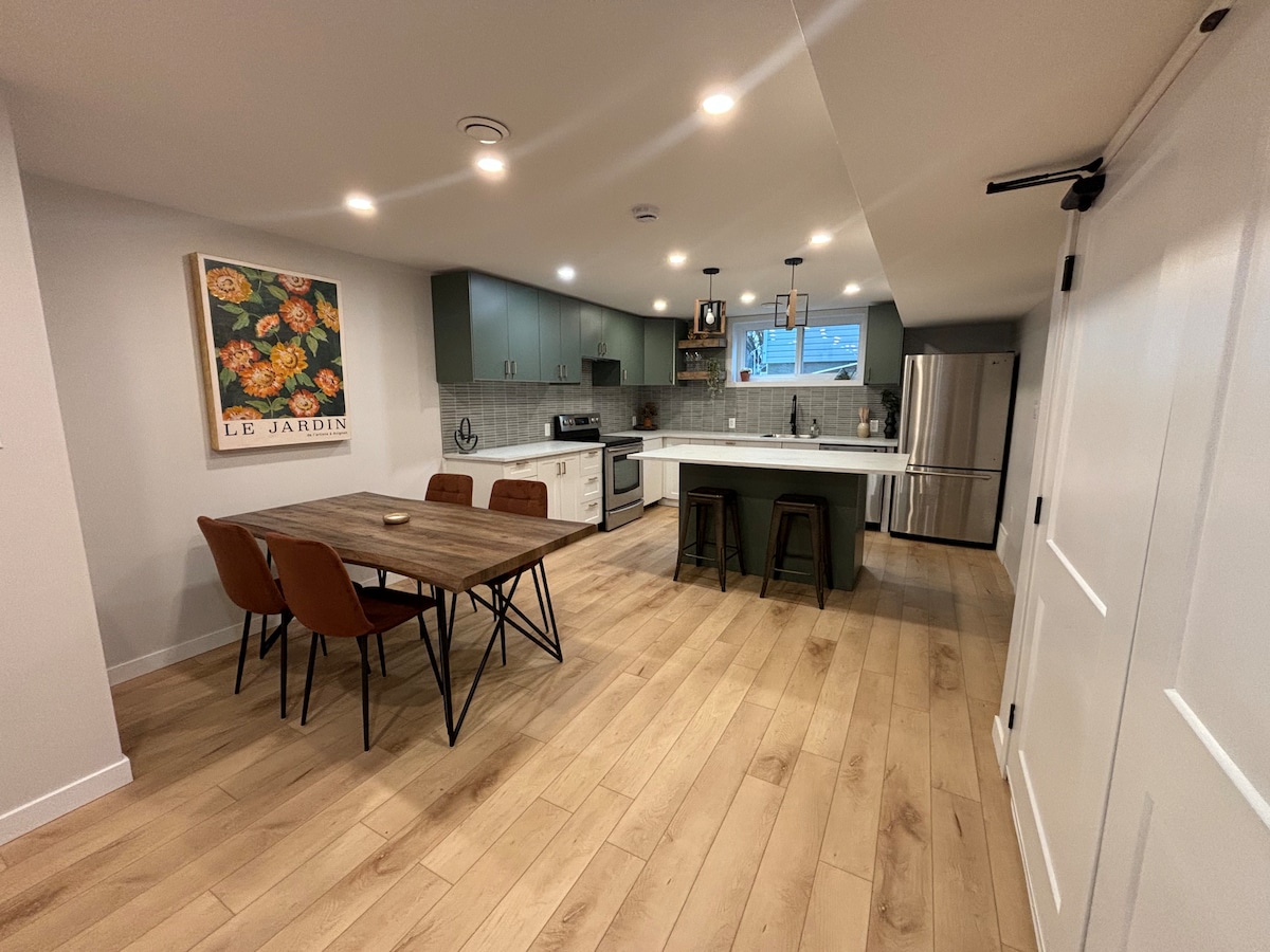 A well-lit kitchen and dining area feature modern cabinets in muted green, complemented by a wooden dining table surrounded by four brown chairs. Stainless steel appliances are visible, with light-colored countertops providing ample workspace. A wall art piece adds a subtle decorative touch.