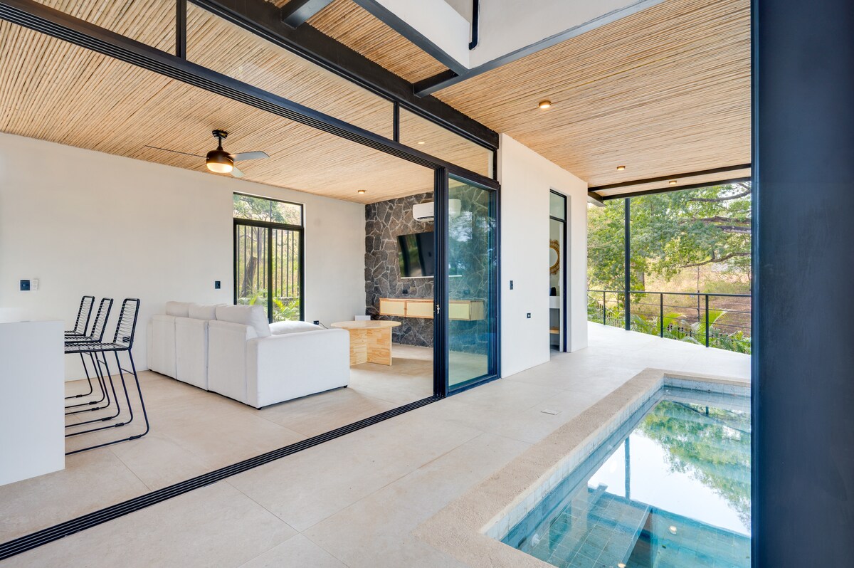 An open-concept living area merges seamlessly with an outdoor space. A plush white sofa faces a mounted TV and wooden coffee table, while large glass doors provide views of the patio and plunge pool. Natural light filters through a wood-paneled ceiling.
