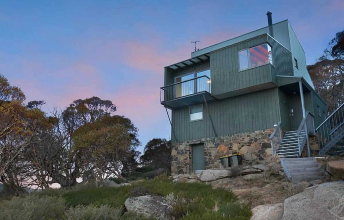 A three-story home with green exterior wooden cladding and a stone base is set amongst trees and rocky terrain. A staircase leads to the entrance, and a balcony is visible on the upper level, allowing scenic views of the surroundings.