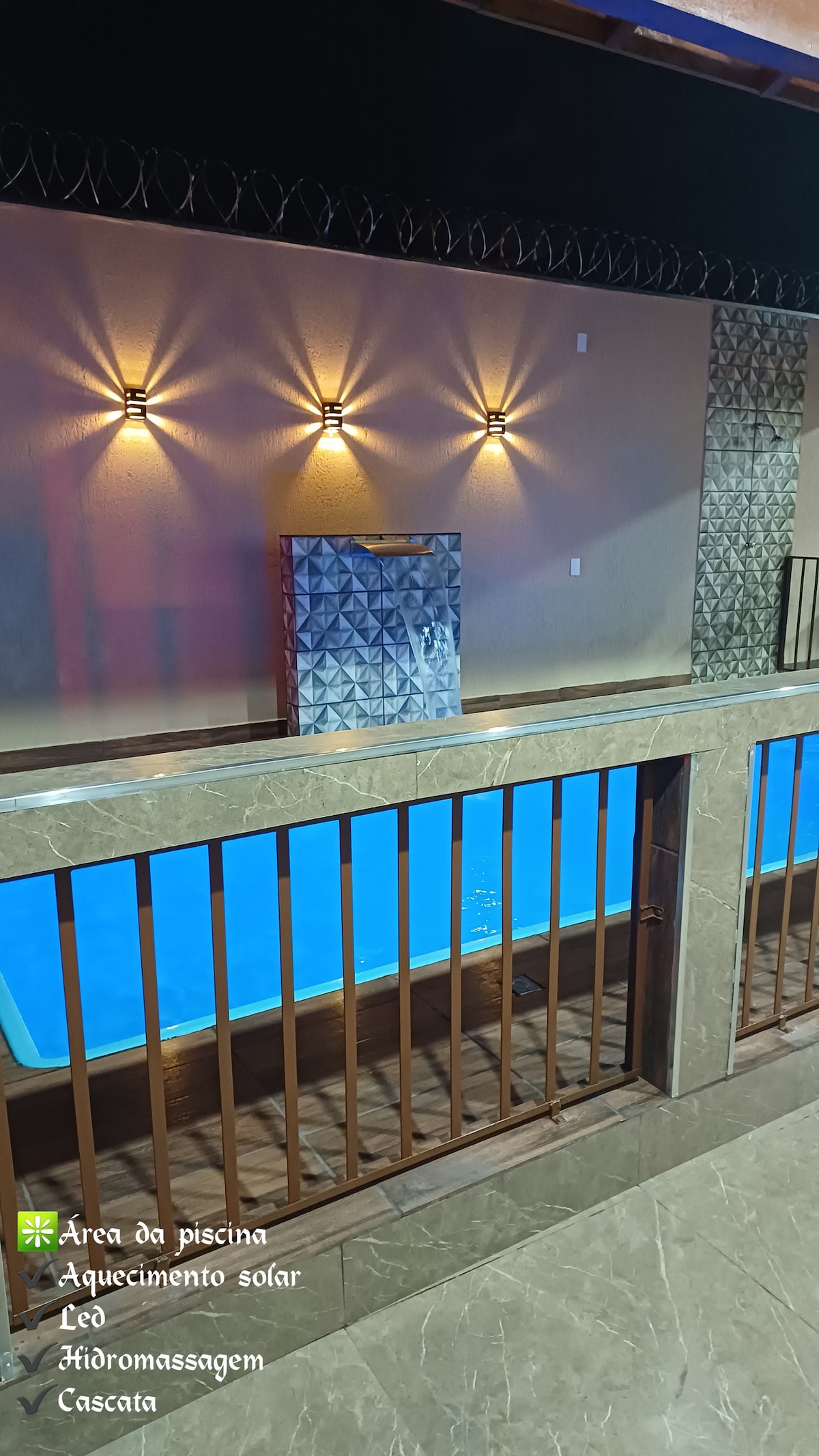 The pool area is complemented by a modern railing and elegant stone finishes. Soft lighting fixtures are mounted on the wall, illuminating the space. The pool features a treatment system, with visible LED lights and a cascading water feature, creating a serene ambiance.