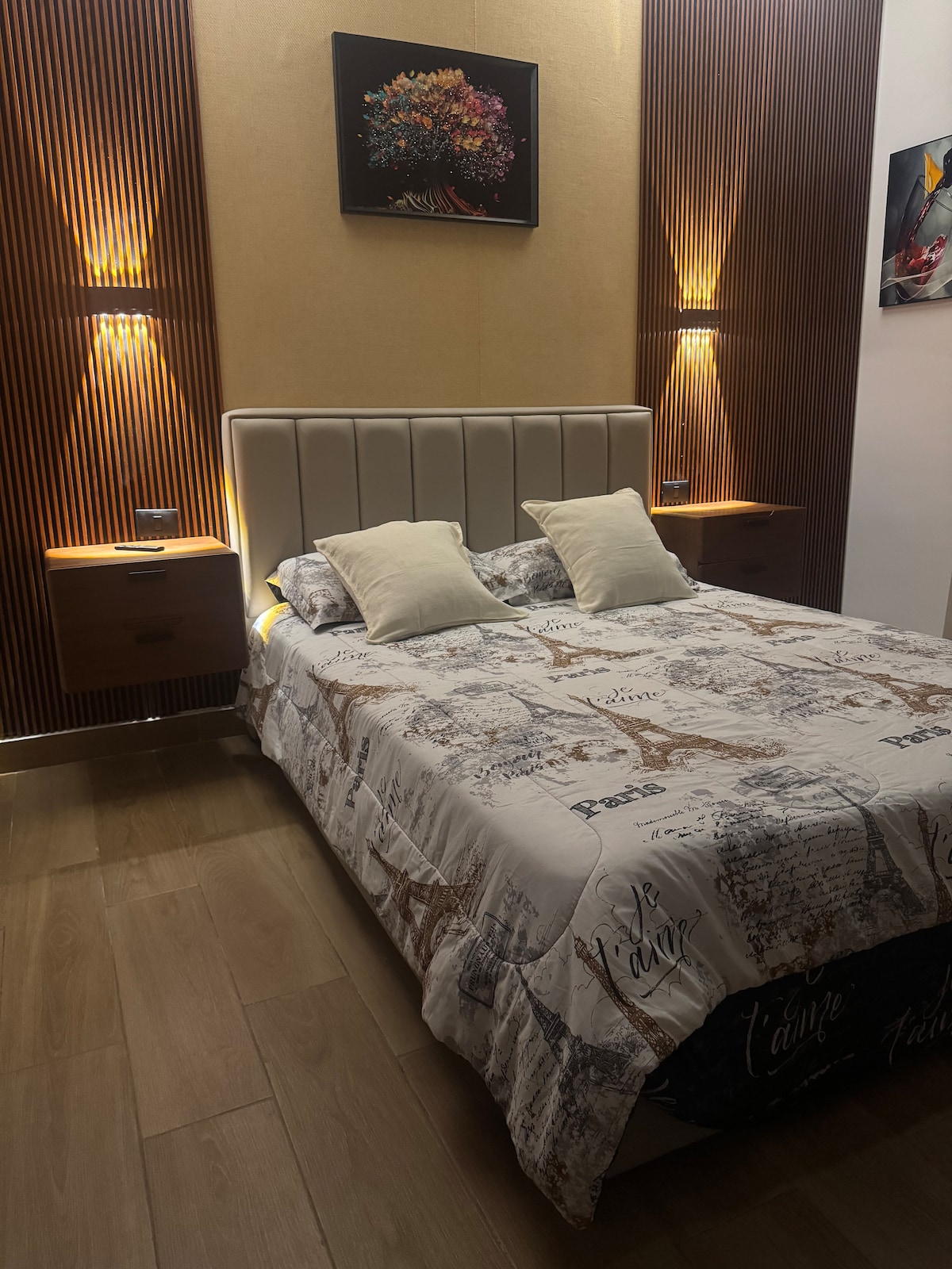 The sleeping area features a padded headboard with integrated lighting on either side. A cozy bed is dressed in a neutral quilt with patterned detailing. Two decorative pillows add to the comfort, while artwork is displayed above the bed, contributing to the room's modern aesthetic.