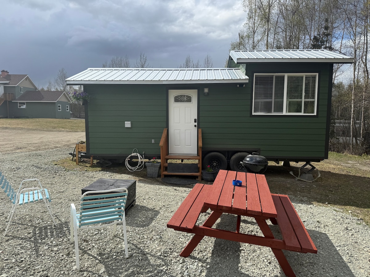 A tiny home features a green exterior with a metal roof and a welcoming front door. A wooden deck leads to the entrance. Outdoor seating includes colorful chairs and a red picnic table, set against a backdrop of grassy space and surrounding trees.