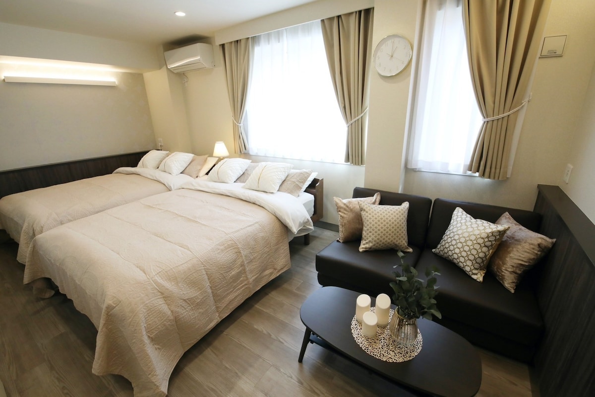 The room features two spacious beds with neutral-toned bedspreads placed against the wall. A comfortable sofa is positioned beside a low coffee table, adorned with decorative pillows and a plant. Natural light filters through the soft curtains of the window, and a wall clock adds a functional touch.