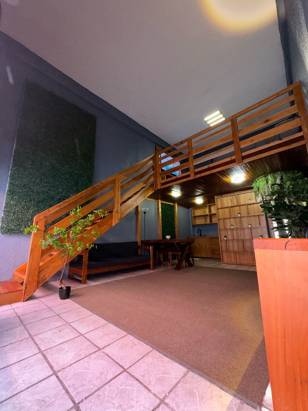 A wooden staircase ascends to a lofted area, illuminated by soft lighting. The spacious living area features a comfortable seating arrangement and a wooden table. Green accents provided by wall plants add a touch of nature to the neutral-toned space.