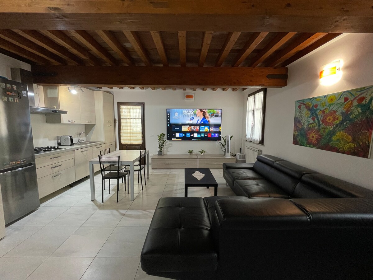An open-plan living area features modern furnishings, including a large black leather sofa and a sleek dining table with four chairs. A wall-mounted Smart TV is positioned above a minimalist cabinet, while natural light enters through windows adorned with light curtains.