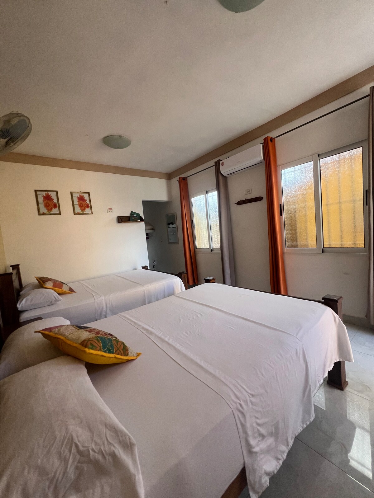 A well-lit room features two spacious beds with white linens and colorful decorative pillows. Natural light enters through large windows adorned with orange curtains, enhancing the clean and modern atmosphere. A fan and air conditioning unit are visible, providing comfort.