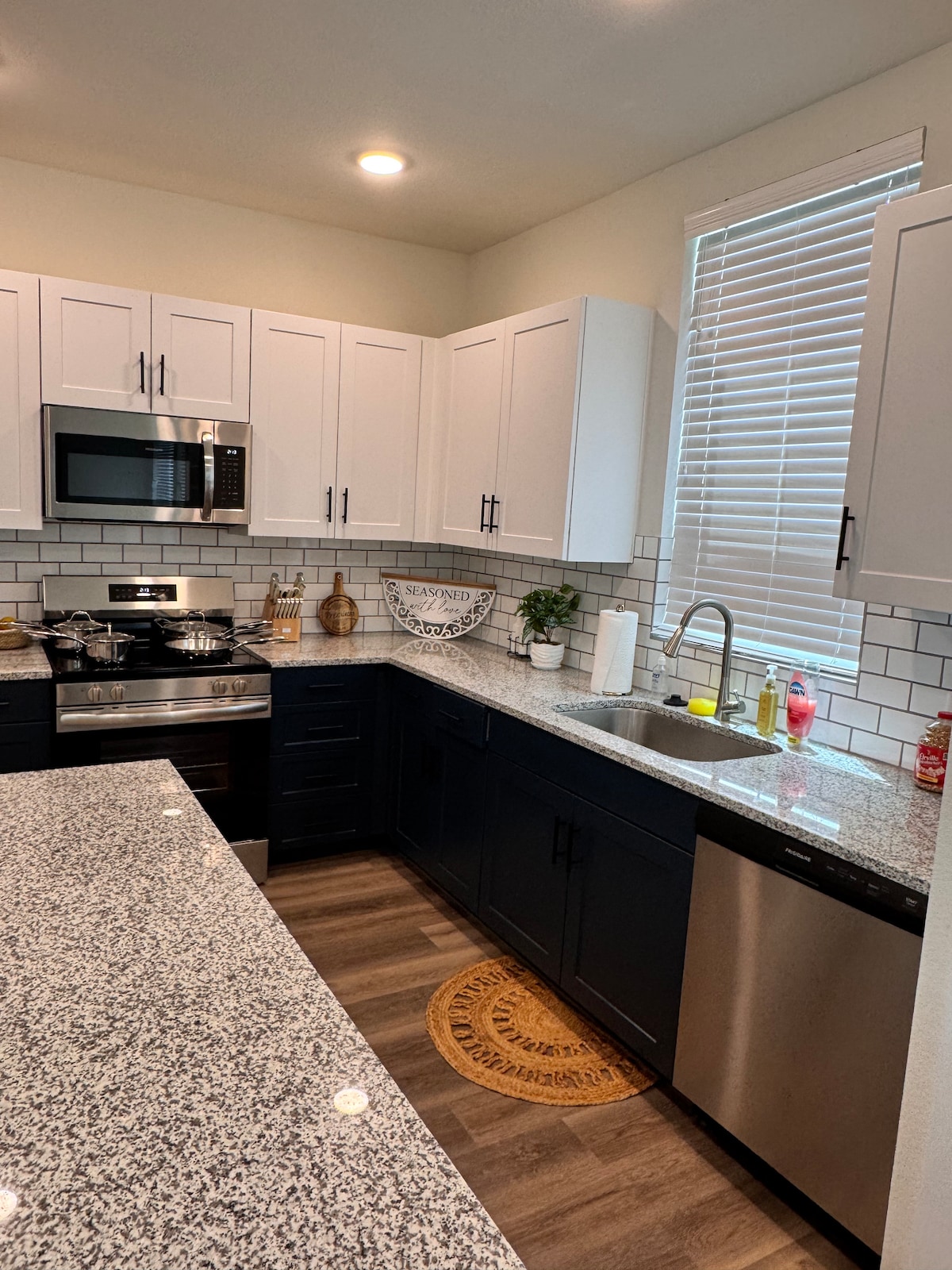 A modern kitchen is showcased with sleek white cabinetry and dark blue accents. Stainless steel appliances are positioned for convenience, while a spacious granite countertop offers ample preparation space. Natural light filters through a window adorned with horizontal blinds, illuminating the stylish tile backsplash.