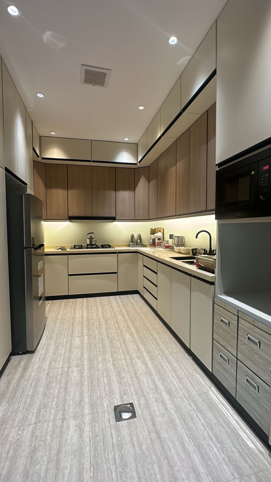A modern kitchen is characterized by sleek cabinetry in neutral tones and stylish wooden accents. Stainless steel appliances, including a refrigerator and built-in microwave, are featured. A streamlined layout provides ample counter space and storage, with soft lighting enhancing the functional environment.