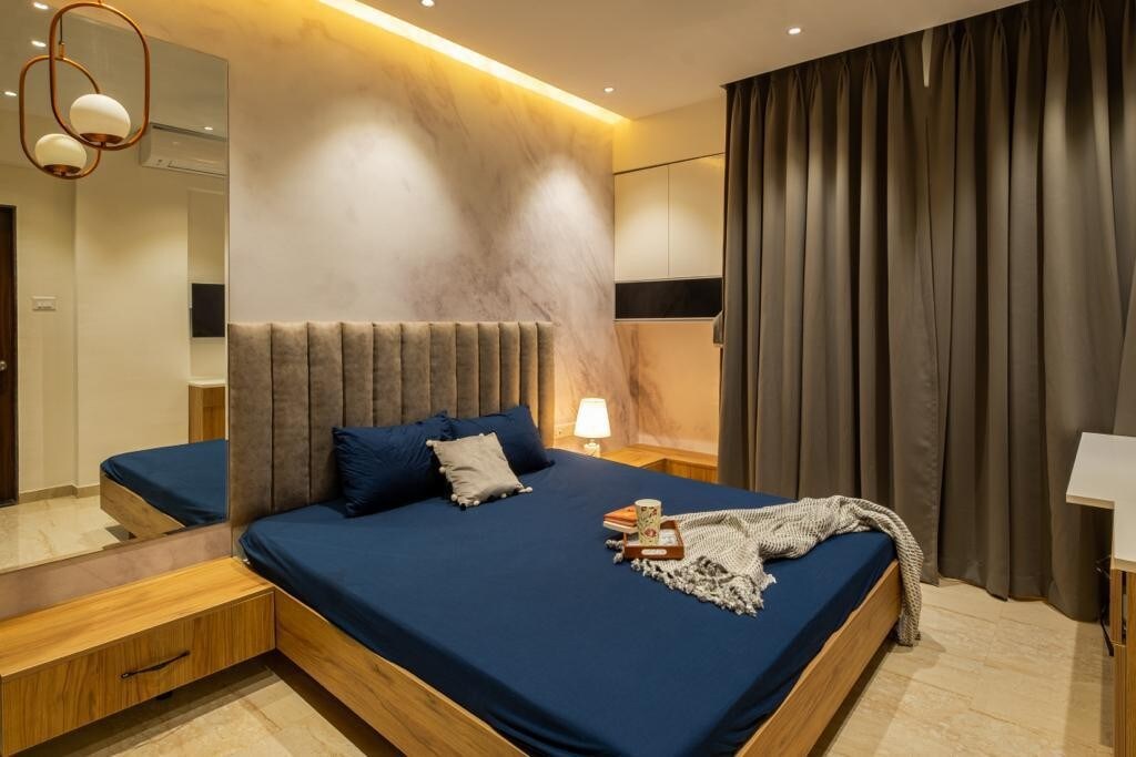 A spacious sleeping area is featured, with a comfortable upholstered headboard and a deep blue bedspread. A decorative pillow lies on the bed, complemented by a soft throw blanket. Warm light is provided by a stylish lamp, and dark curtains frame the window.