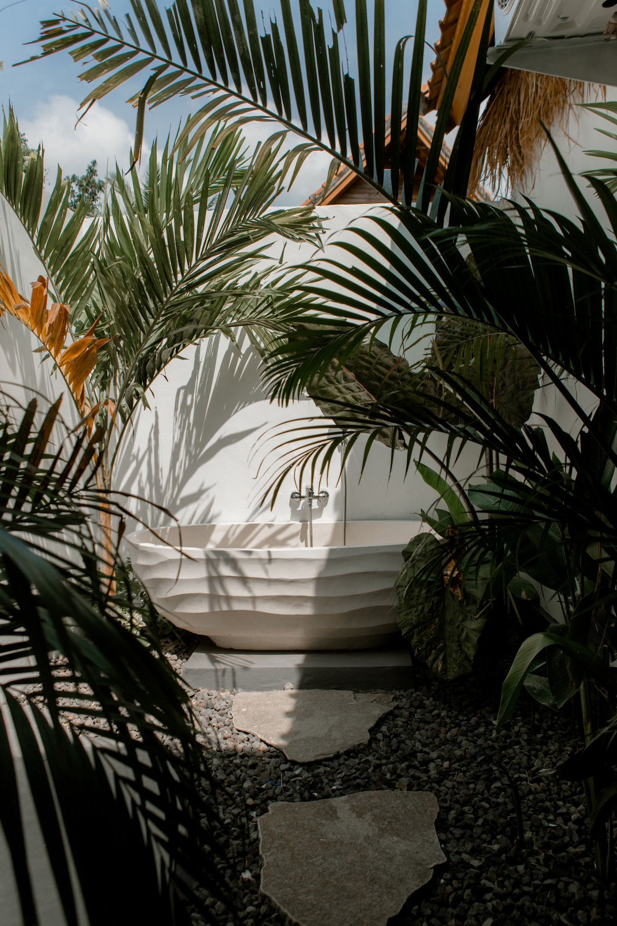 A tranquil outdoor bathroom is framed by lush greenery and tall palm leaves, creating a serene atmosphere. A softly contoured bathtub is positioned on a gravel pathway, with smooth stone tiles leading the way. Light filters through the foliage, enhancing the secluded feel.