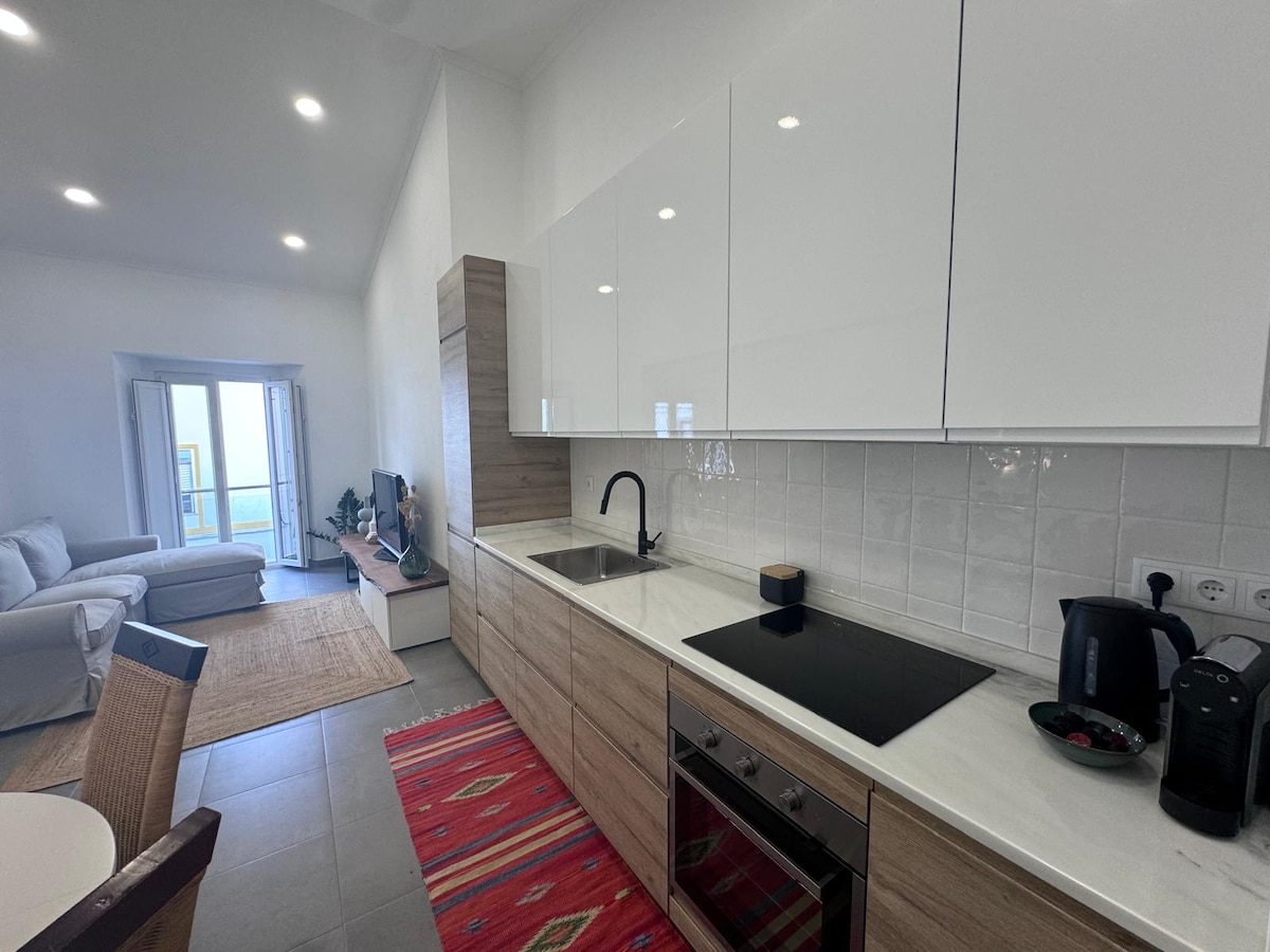 A modern kitchen features sleek white cabinets with wood accents, showcasing a clean, minimalistic design. A black cooktop and an electric kettle are visible on the countertop. A cozy dining area is set nearby, with a natural fiber rug enhancing the space's warmth.