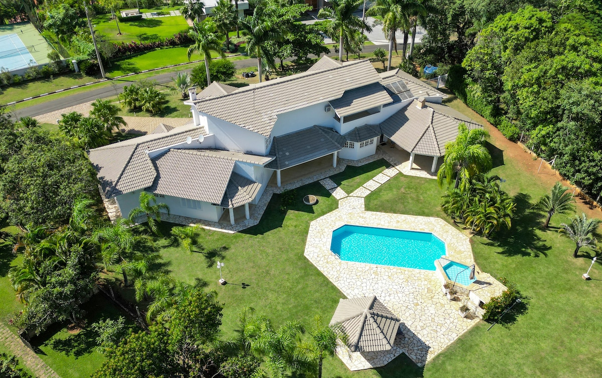 The aerial view showcases a spacious property surrounded by lush greenery. A glistening pool is positioned at the center, accompanied by lounge chairs. Multiple covered gazebos are visible in the well-manicured lawn, providing inviting outdoor spaces for relaxation and leisure.