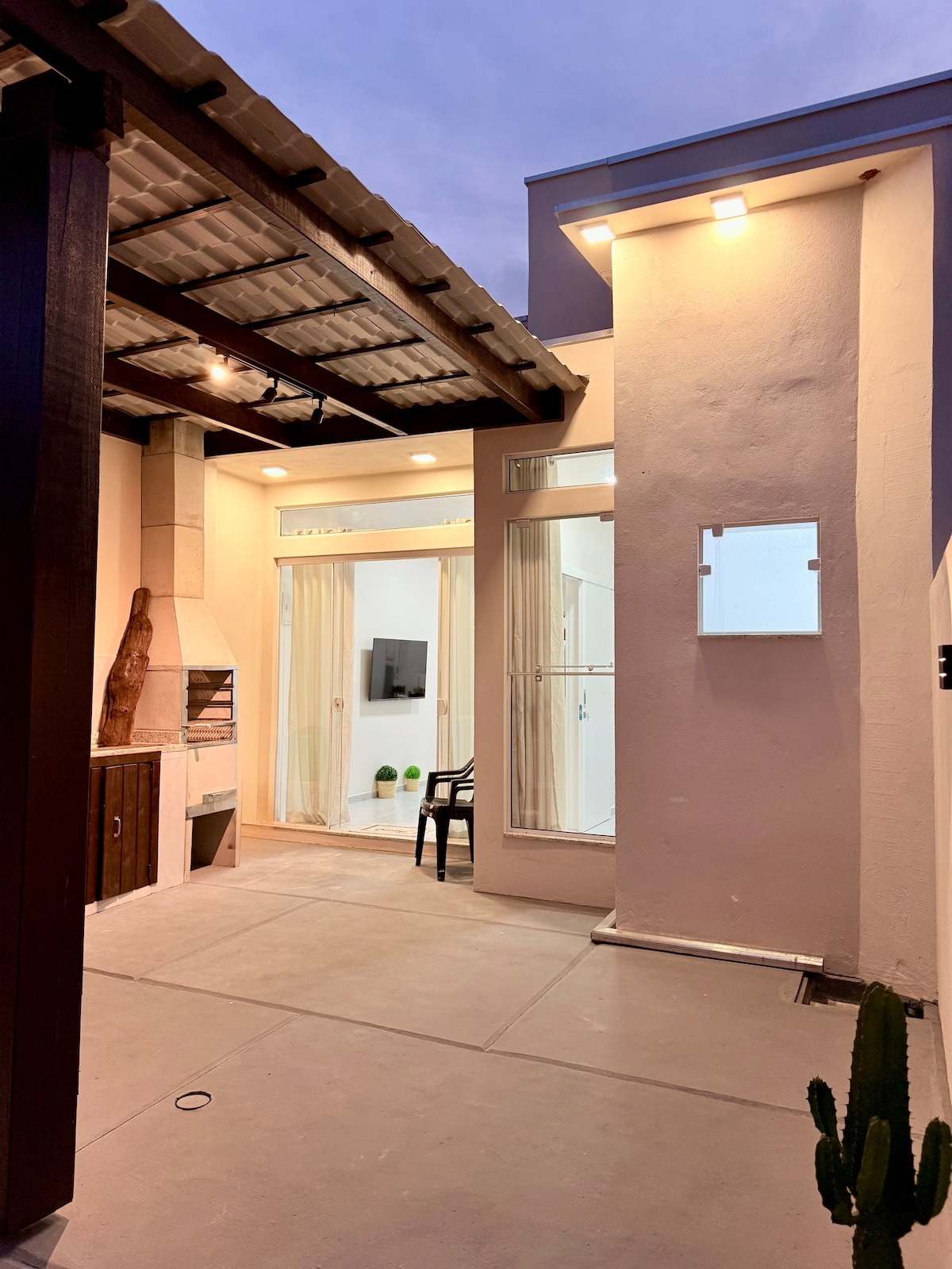 An outdoor area is presented, featuring a barbecue grill and a black chair positioned on a light-colored patio. Tall walls enclose the space, with large windows allowing natural light to enter. Soft lighting is provided by fixtures mounted on the overhead structure, enhancing the ambiance.