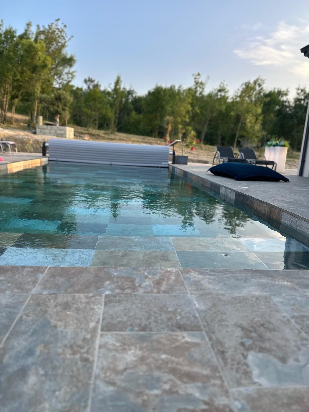 A calm swimming pool is surrounded by a tiled deck and lush greenery. The pool features a retractable cover, and lounge chairs are positioned nearby, inviting relaxation. The serene landscape complements the tranquil atmosphere, making it an ideal outdoor space.