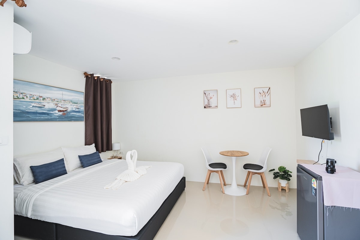 A bright room features a spacious bed with white linens, accented by dark pillows. A small dining table with two chairs is set against the wall. Artwork adorns the wall above the bed, while a television is mounted opposite, providing entertainment options.
