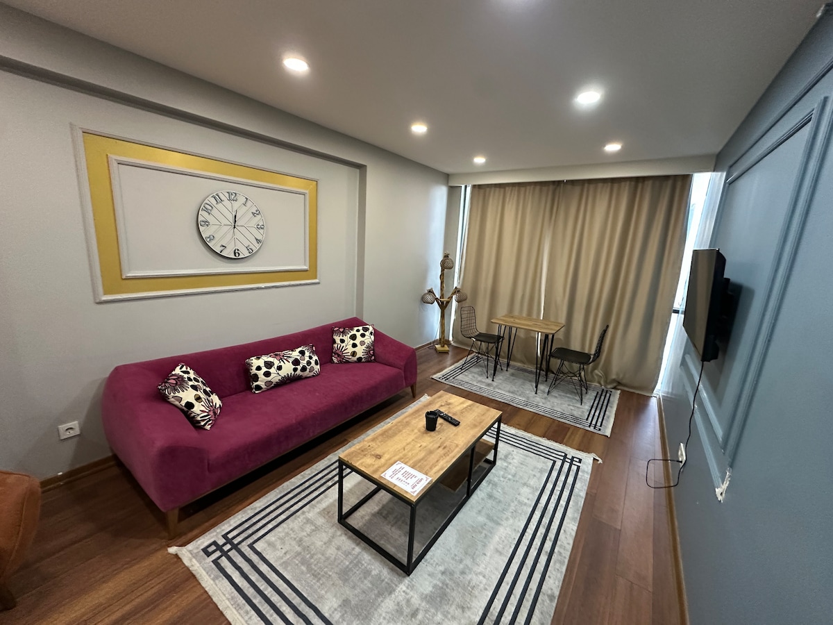 A modern living space features a vibrant purple sofa with decorative pillows, complimented by a wooden coffee table. A clock and wall art create visual interest. A dining area with two chairs is present, along with large windows draped with soft curtains that allow natural light to fill the room.