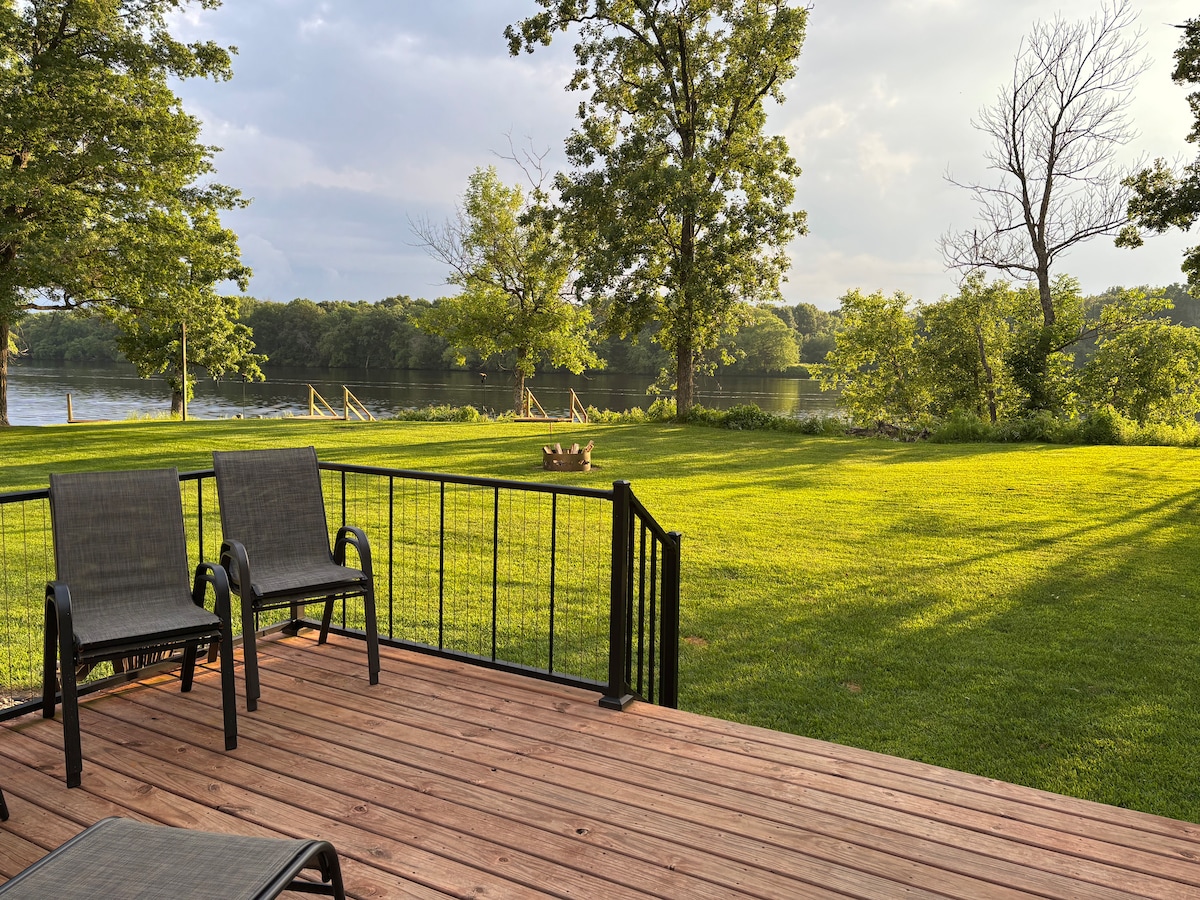 A wooden deck features two chairs positioned for viewing the expansive green lawn. Tall trees frame the scene, which overlooks the tranquil Wisconsin River. A fire pit is visible in the background, enhancing the outdoor setting.