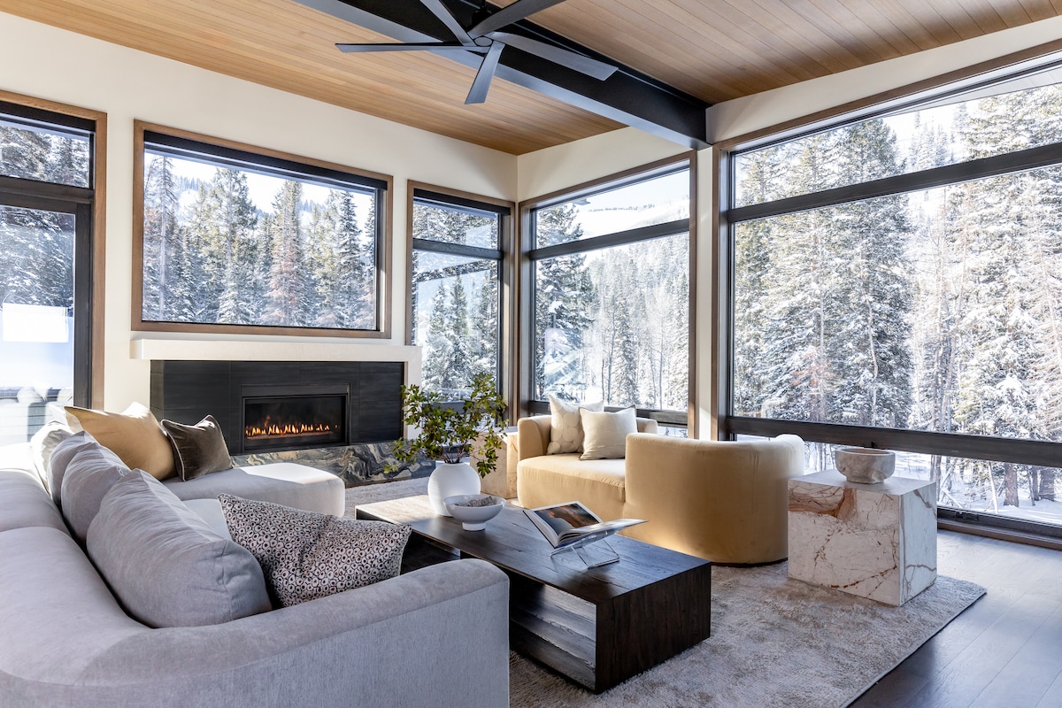 A spacious living area is highlighted by large windows, offering a view of the snowy outdoors. A cozy sectional sofa and two accent chairs surround a wooden coffee table. A sleek fireplace is positioned on the wall, enhancing the comfortable atmosphere of the room.