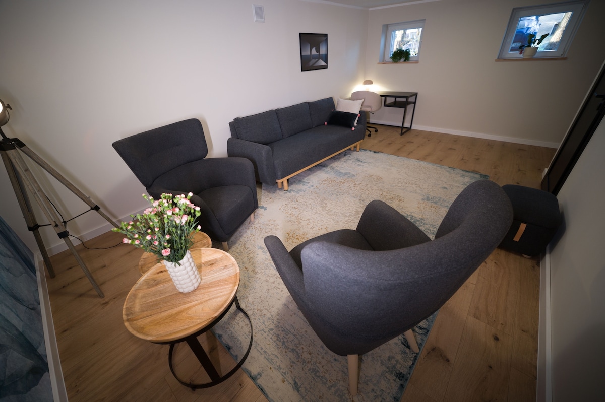 A comfortable living area showcases a gray sofa paired with two matching armchairs on a light wooden floor. A round wooden coffee table is positioned at the center, adorned with a small vase of flowers. Natural light enters through the nearby window, illuminating the space.