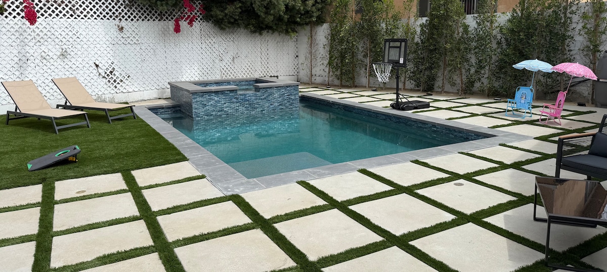 An outdoor space features a modern swimming pool surrounded by a stone patio. A spa area is integrated within the pool. Loungers are distributed on the grass-like area, with a small basketball hoop and colorful seating for children nearby.