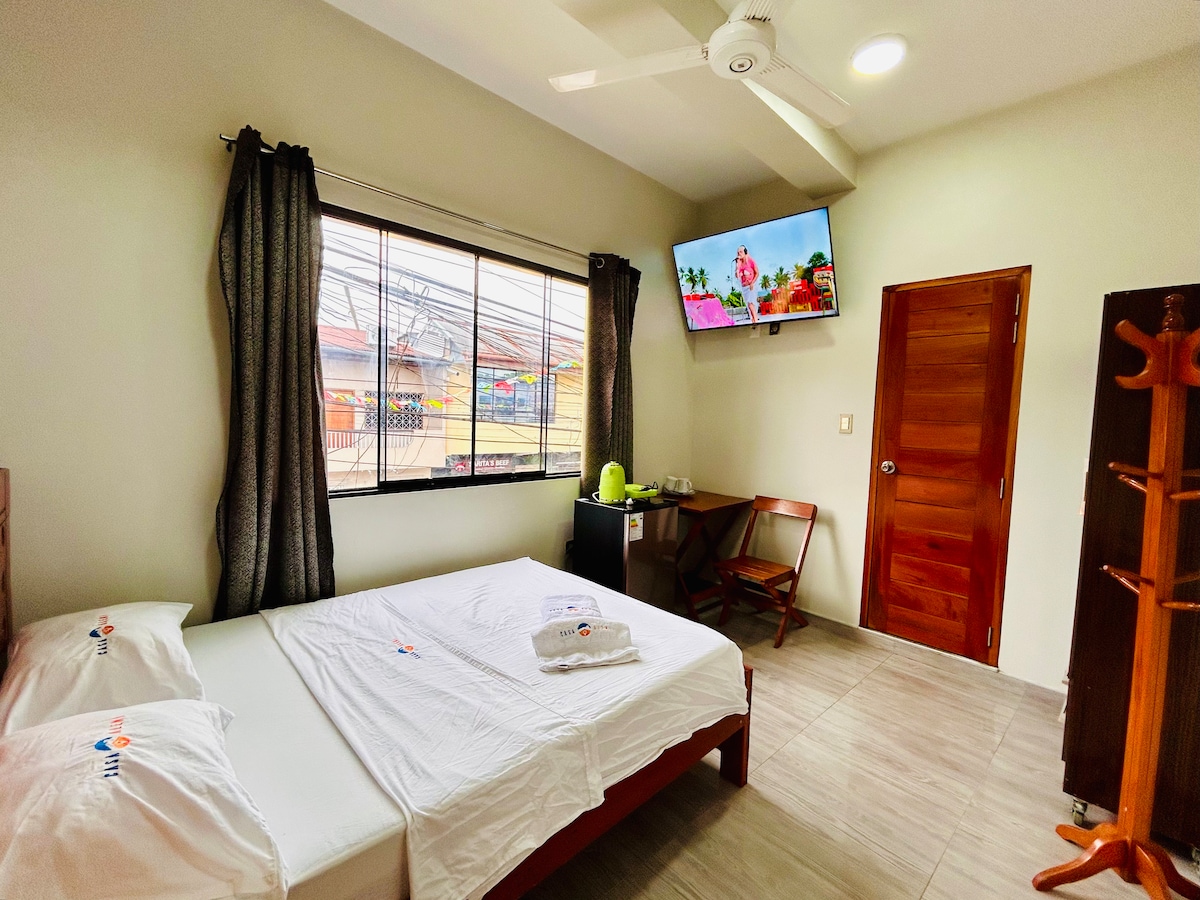 A comfortable room is presented with a spacious bed, accompanied by two neatly arranged towels. A flat-screen smart TV is mounted on the wall, and a mini-fridge is positioned beside a small table and chair near a large window dressed with dark curtains.