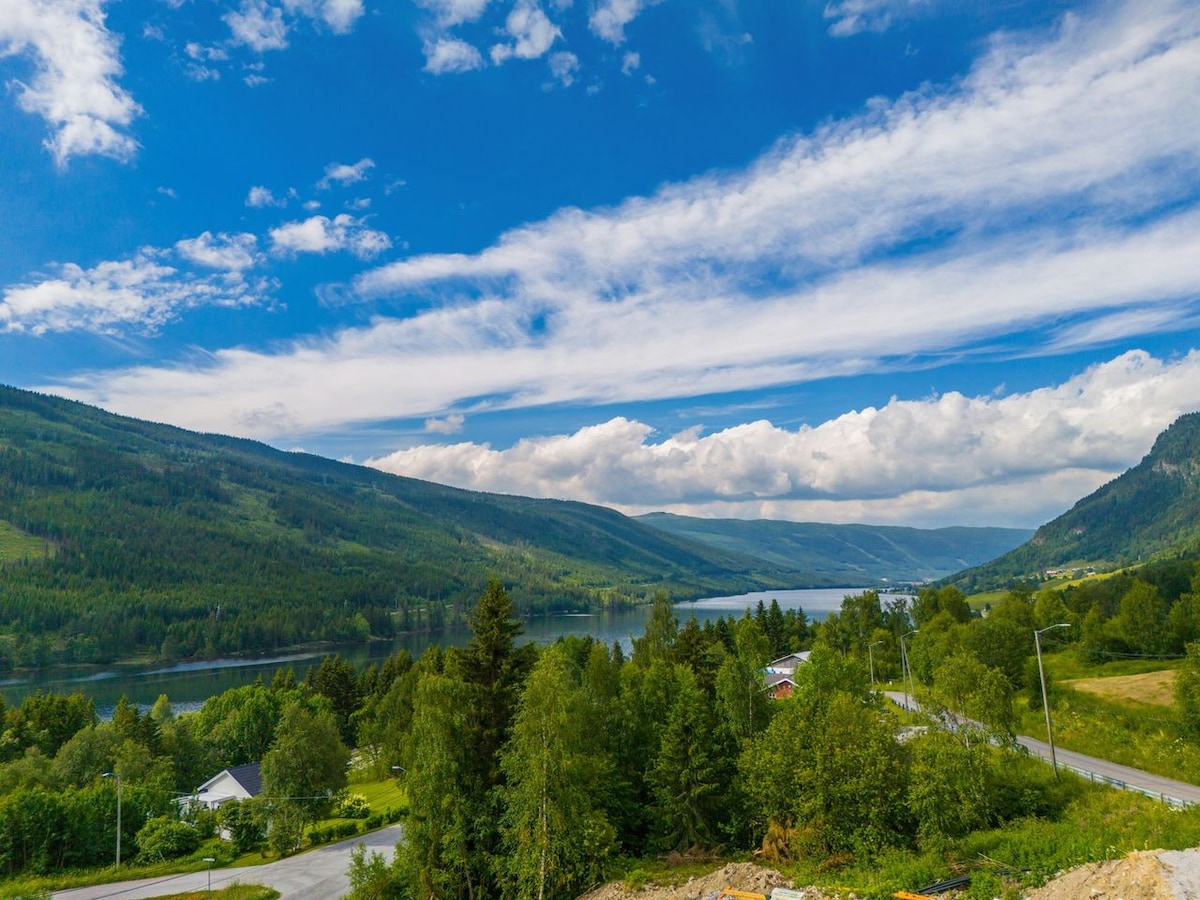 A scenic landscape features rolling green hills and a wide river, reflecting the blue sky above. Fluffy white clouds drift across the horizon. The view includes scattered trees and a small road, providing context to the natural surroundings.