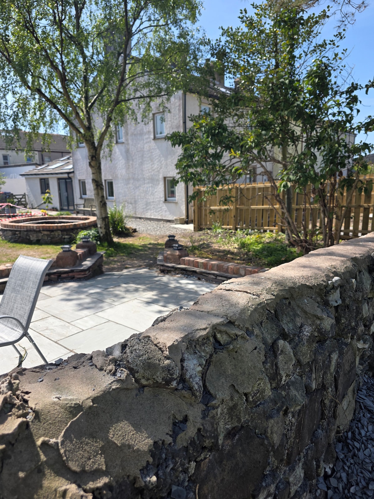A private outdoor area is shown with a stone wall in the foreground. Beyond the wall, a well-maintained patio is visible, surrounded by various plants and a tree. The background features a two-story cottage with multiple windows and a fenced area, providing a peaceful garden setting.