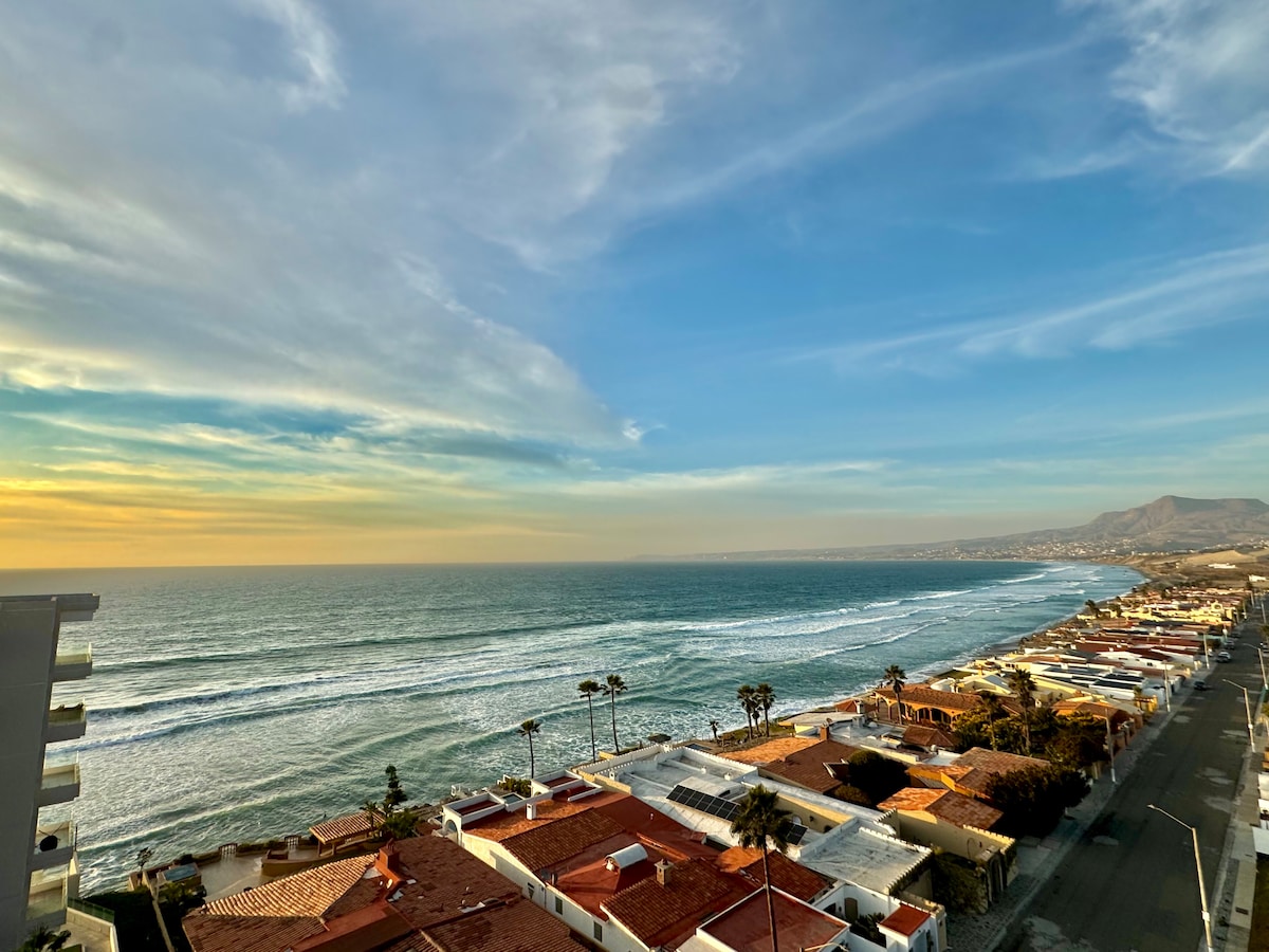 An expansive view of the coastline captures gentle waves lapping against the shore. Colorful rooftops of nearby structures create a vibrant foreground, while the horizon blends with the sky, showcasing hues of blue and warm sunlight.