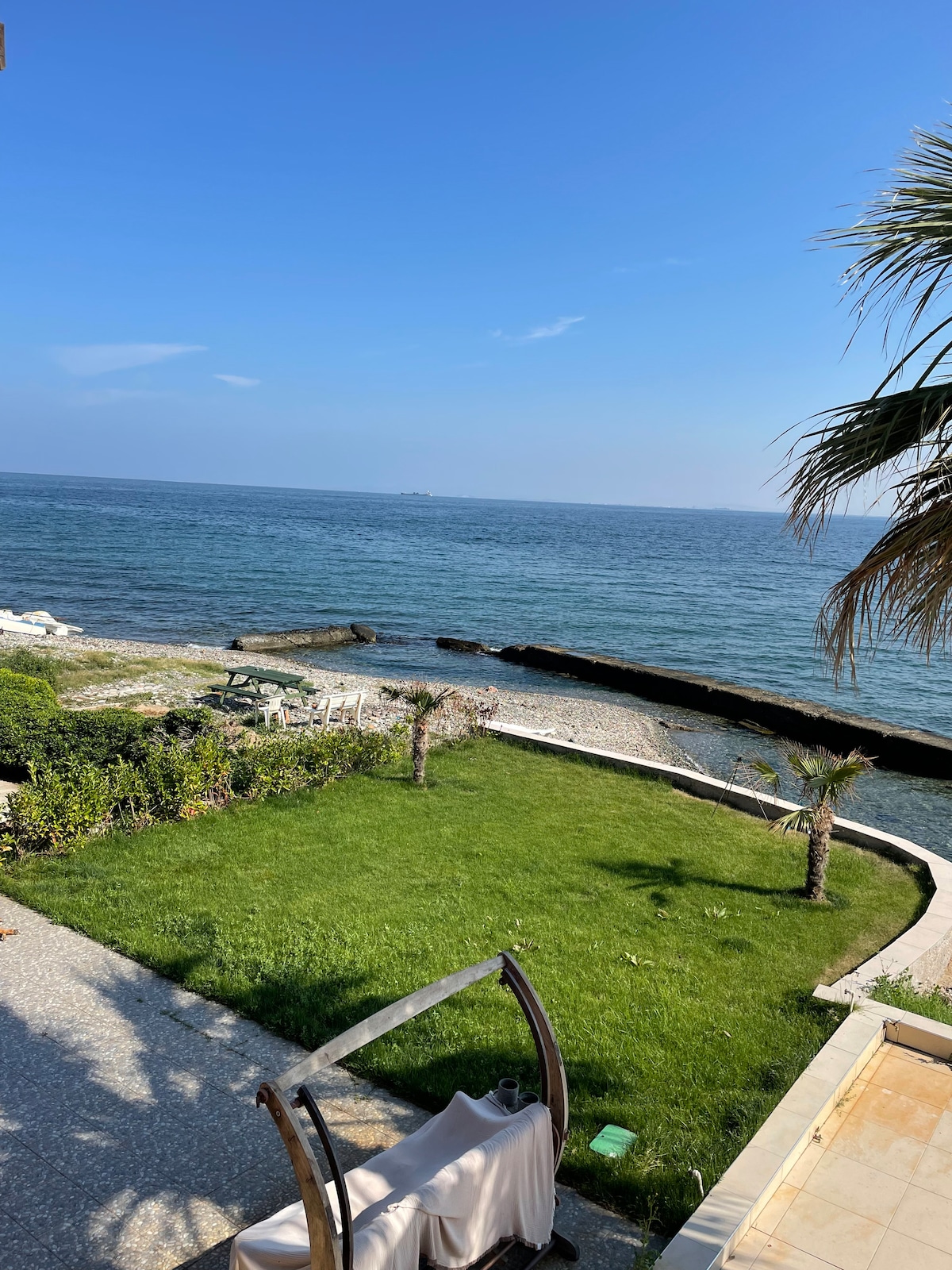 A serene oceanfront view features gently lapping waves against rocky shoreline. Lush green grass leads towards the water, bordered by palm trees and stones. Clear blue skies hold the promise of a sunny day, enhancing the natural coastal landscape.