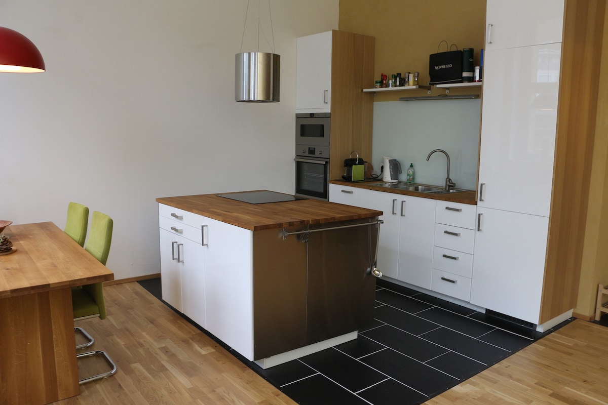 A modern kitchen area showcases a central island with a wooden countertop and stainless steel features. Clean white cabinets are complemented by wooden accents. An oven and microwave are built into the wall, with a dining table nearby and green chairs enhancing the dining space.