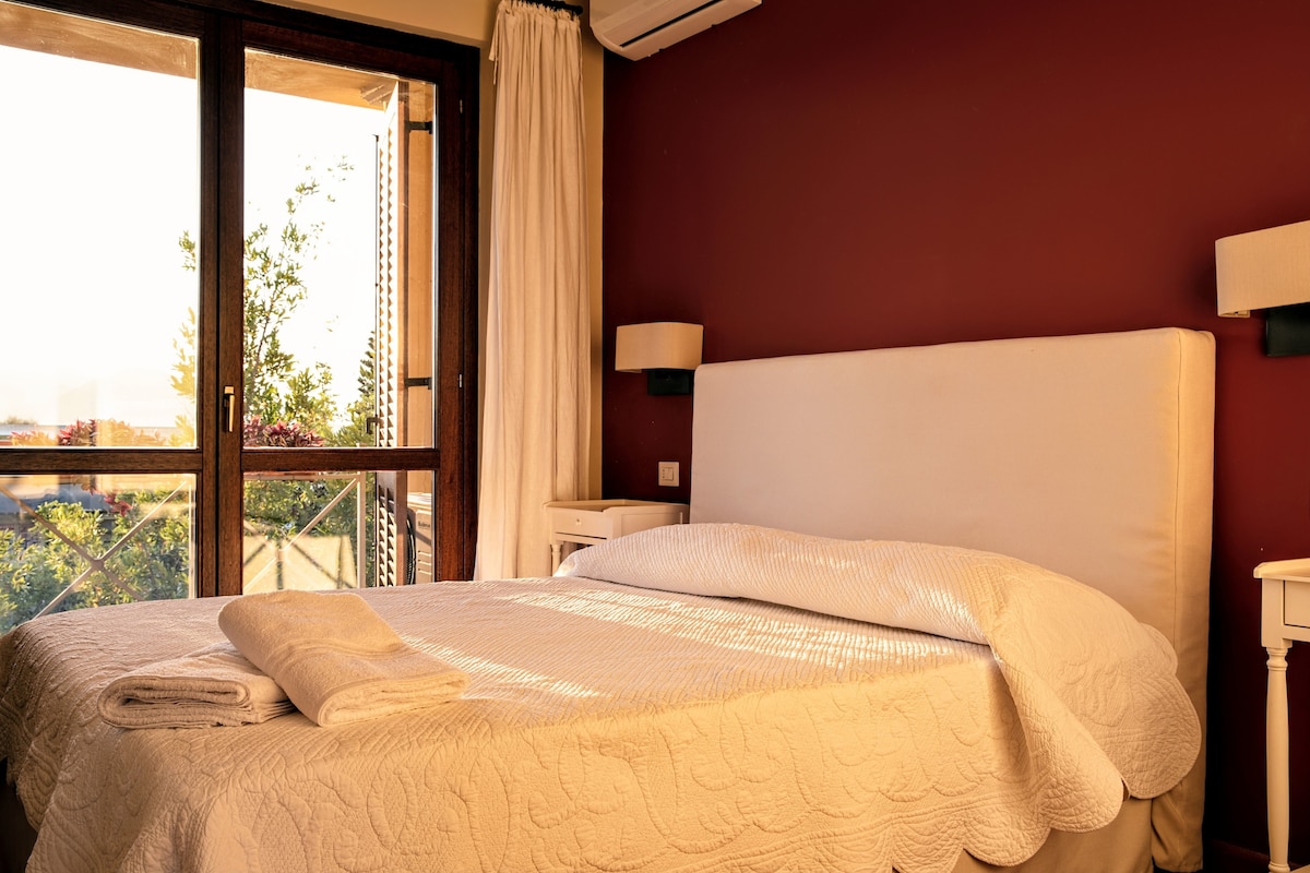 A bright bedroom features a spacious bed covered with a textured white quilt. Two bedside tables with lamps flank the bed, and large windows allow natural light to illuminate the room. Soft curtains frame the windows, hinting at the greenery outside.