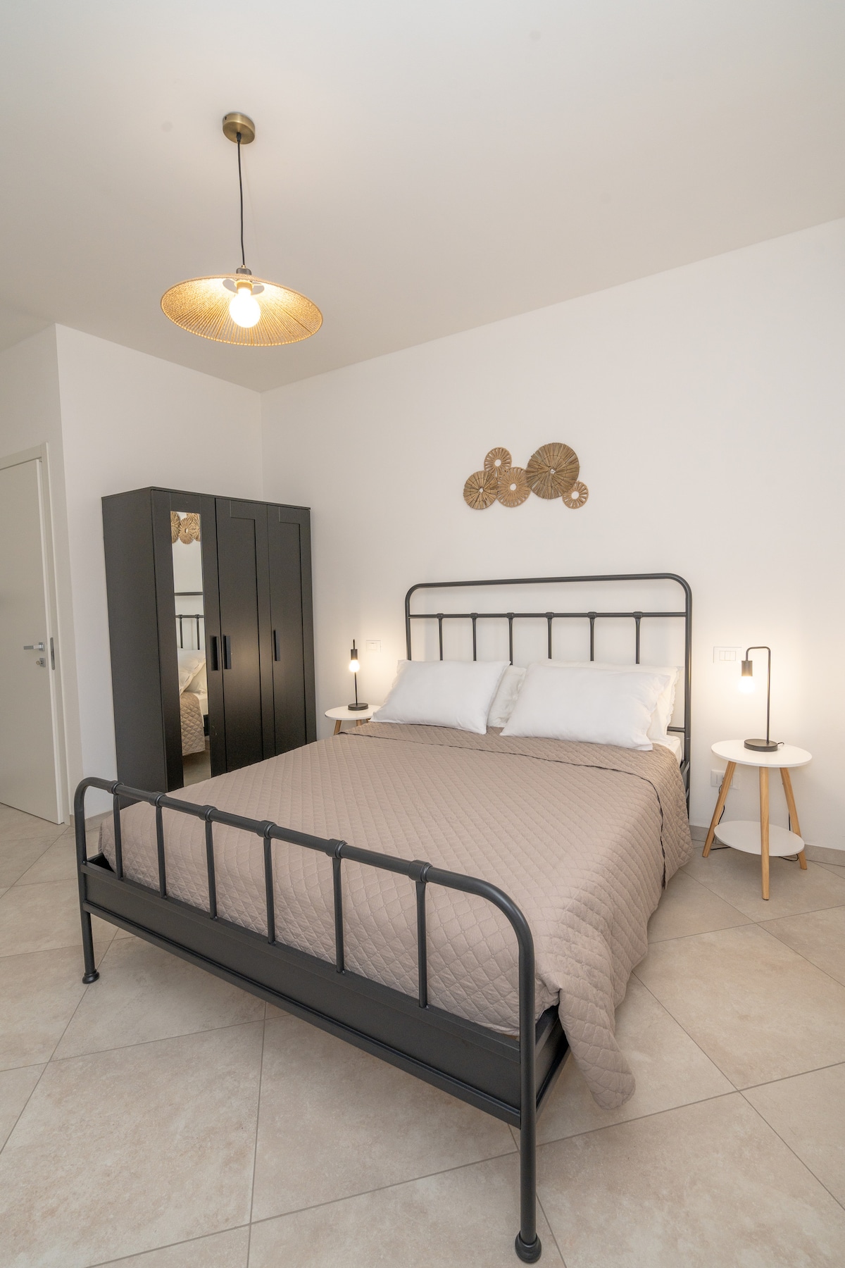 The bedroom features a metal bed frame with a neutral quilt and two comfortable pillows. Nightstands with lamps are positioned on either side of the bed. A wardrobe is included against the wall, and decorative wall accents provide a simple touch.