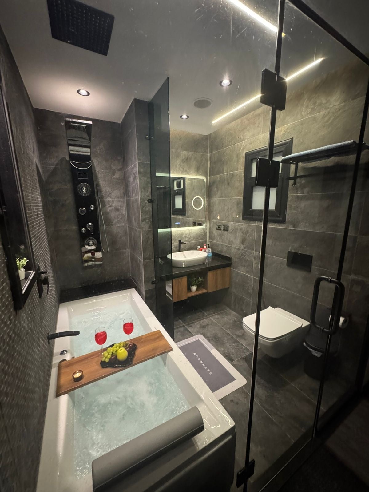 The modern bathroom is equipped with a glass-enclosed hot Jacuzzi tub fitted with a wooden bath tray displaying drinks and decorative items. A sleek vanity with a round mirror is positioned above the sink, alongside a wall-mounted toilet and towel rack. Soft lighting complements the dark tile accents.