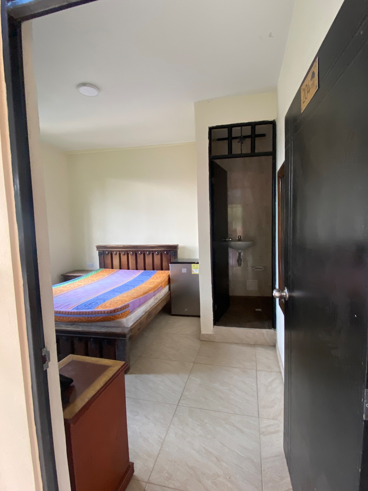 A simple room is showcased with a spacious bed covered in a colorful striped blanket. A small nightstand and a refrigerator are positioned nearby. A private bathroom area is visible in the corner, accessible through an open doorway.