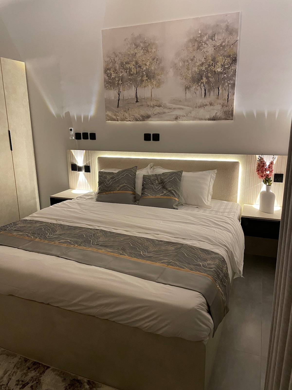 A master bedroom is highlighted by a spacious bed adorned with decorative pillows and a stylish headboard. Nightstands with soft lighting are positioned on either side. A framed artwork featuring trees hangs above the bed, enhancing the serene ambiance of the room.