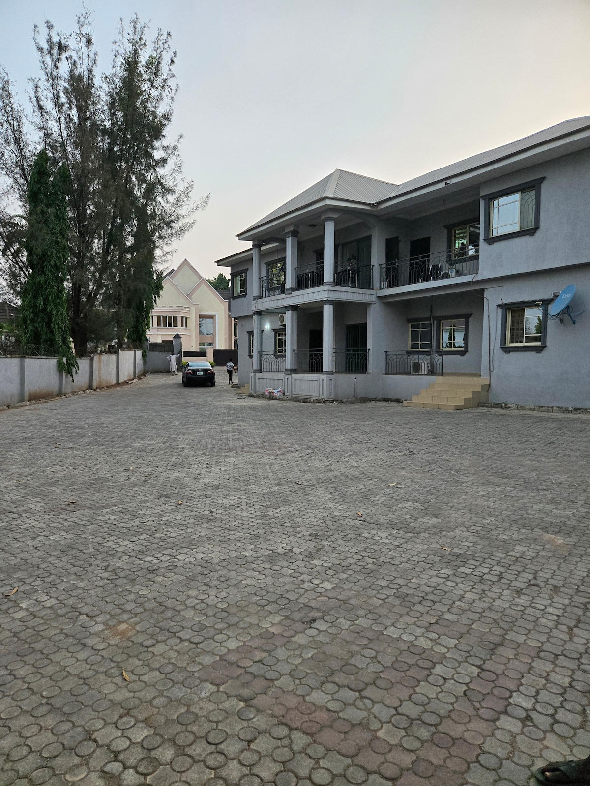 A spacious exterior view captures the two-story condo set within a gated area. The building features a covered porch with multiple columns and a symmetrical design. Surrounding greenery adds a natural element, while a stone-paved parking area accommodates vehicles, providing convenient access.