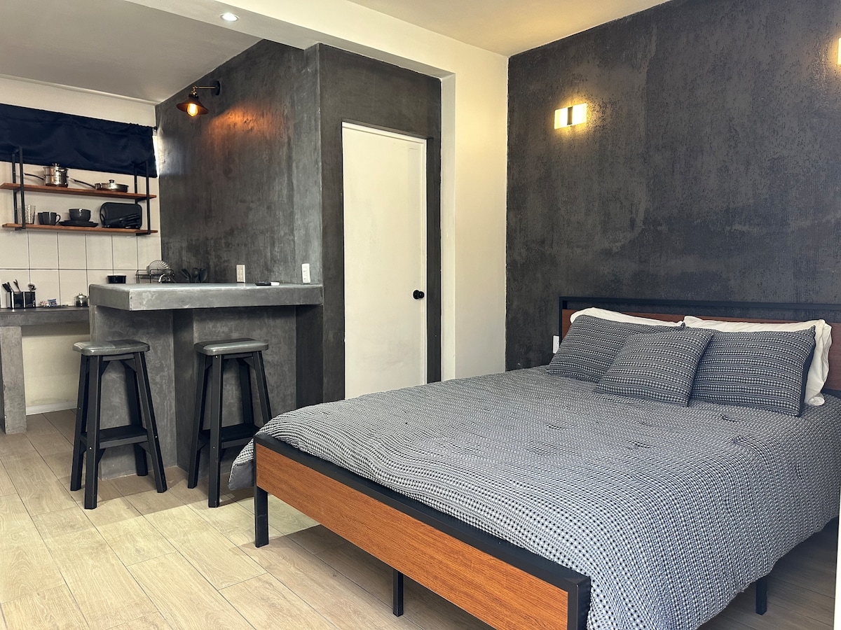A simple yet modern sleeping area features a spacious bed with patterned bedding. A minimalist kitchenette with bar stools is visible in the background, along with open shelving displaying dishware. Neutral-colored walls contribute to a calm atmosphere.