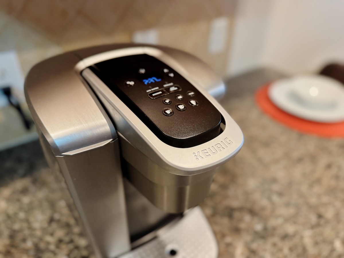 A sleek coffee maker is positioned on a granite countertop, showcasing a modern design with various buttons and a digital display. The surrounding area features a neutral color palette, contributing to a clean and organized kitchen space.