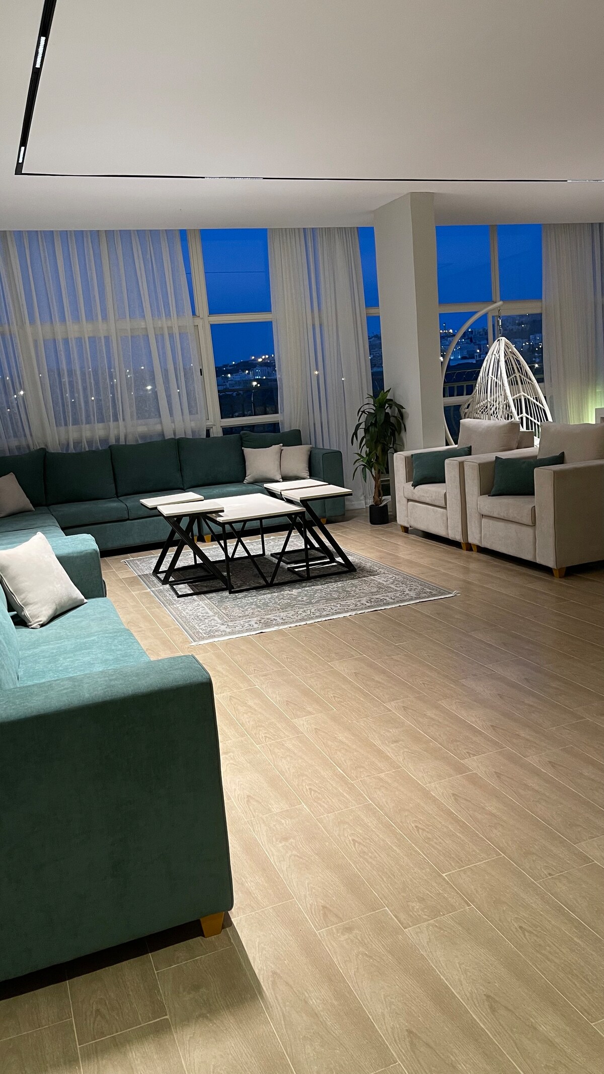 A spacious living area is showcased with a large, L-shaped sofa in soft teal and light-colored accent chairs. A central coffee table is positioned on a patterned rug, complemented by large windows that offer city views and filtered natural light.