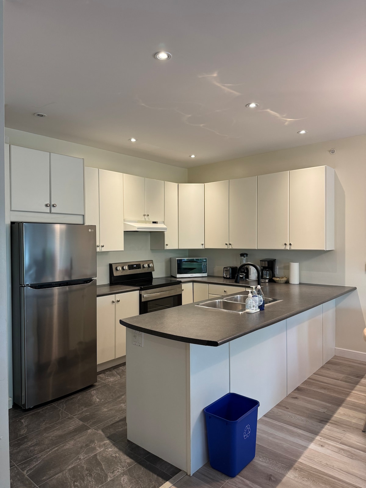 A modern kitchen is equipped with stainless steel appliances, including a refrigerator and stove. Light-colored cabinets provide ample storage, and a dark countertop offers workspace. Recessed lighting illuminates the area, which features a compact table and a blue recycling bin.
