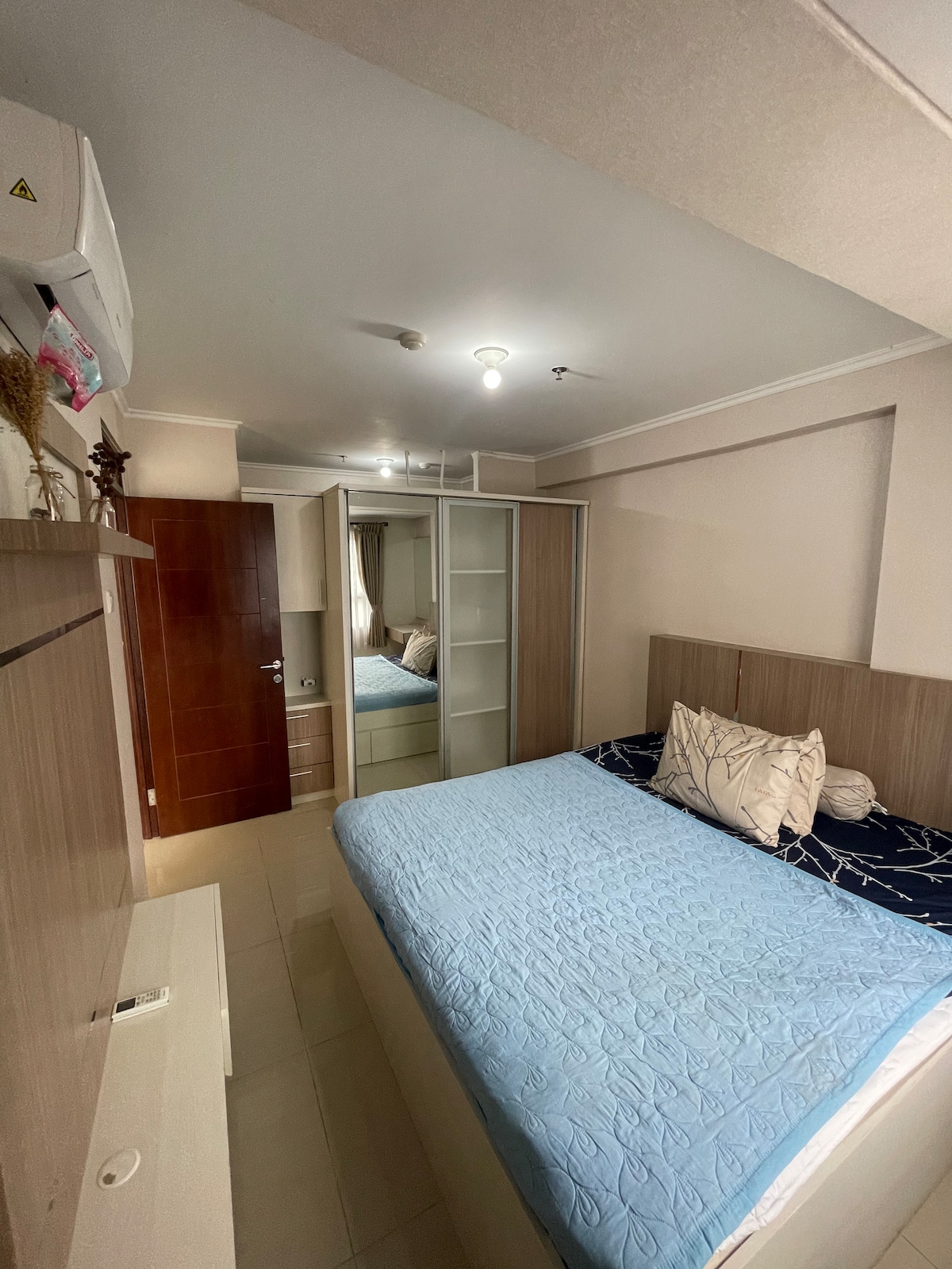 A cozy sleeping area features a spacious bed with a light blue quilt and decorative throw pillows. A large mirror and fitted wardrobes are positioned against the opposite wall, reflecting natural light. Air conditioning unit is visible above, contributing to a comfortable environment.