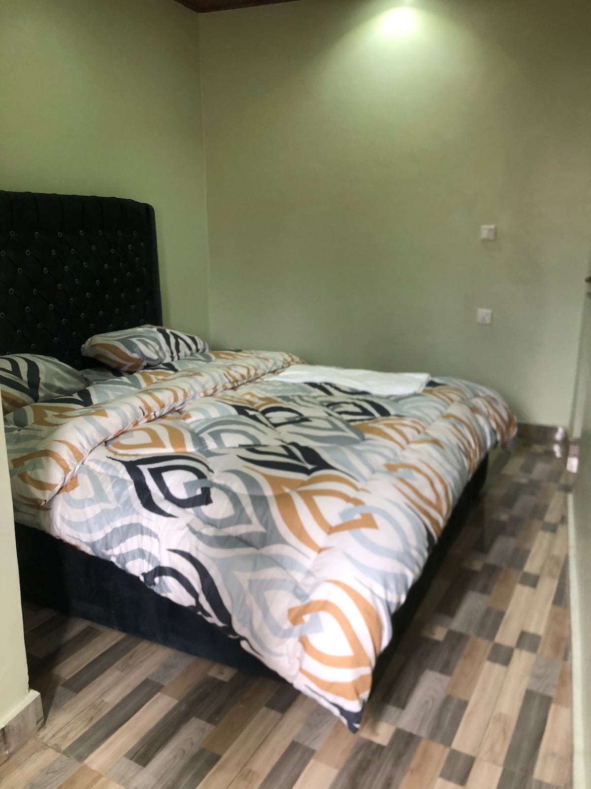 A neatly arranged sleeping area features a dark upholstered headboard and a bed covered with a patterned duvet in neutral tones. Soft lighting highlights the walls, while wooden flooring adds warmth to the ambiance.
