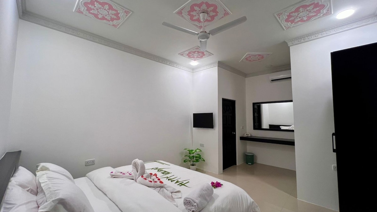 A modern sleeping area features a spacious bed with crisp white linens and decorative towels. The ceiling displays floral designs, while a ceiling fan provides airflow. A minimalist desk area is visible in the background, illuminated by natural light from a nearby window.