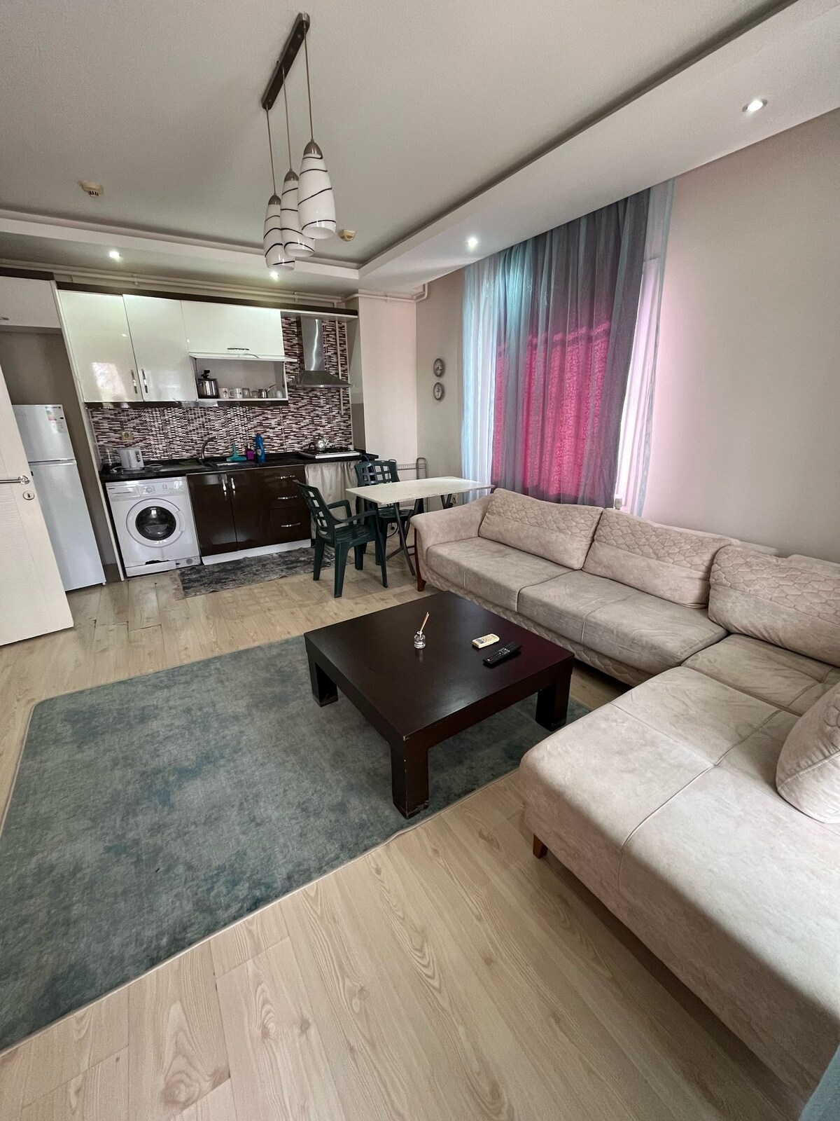 A well-lit living area features a comfortable sectional sofa and a dark wooden coffee table. The kitchenette includes modern appliances and is complemented by decorative tiles. Natural light filters through sheer curtains, adding a soft touch to the space.