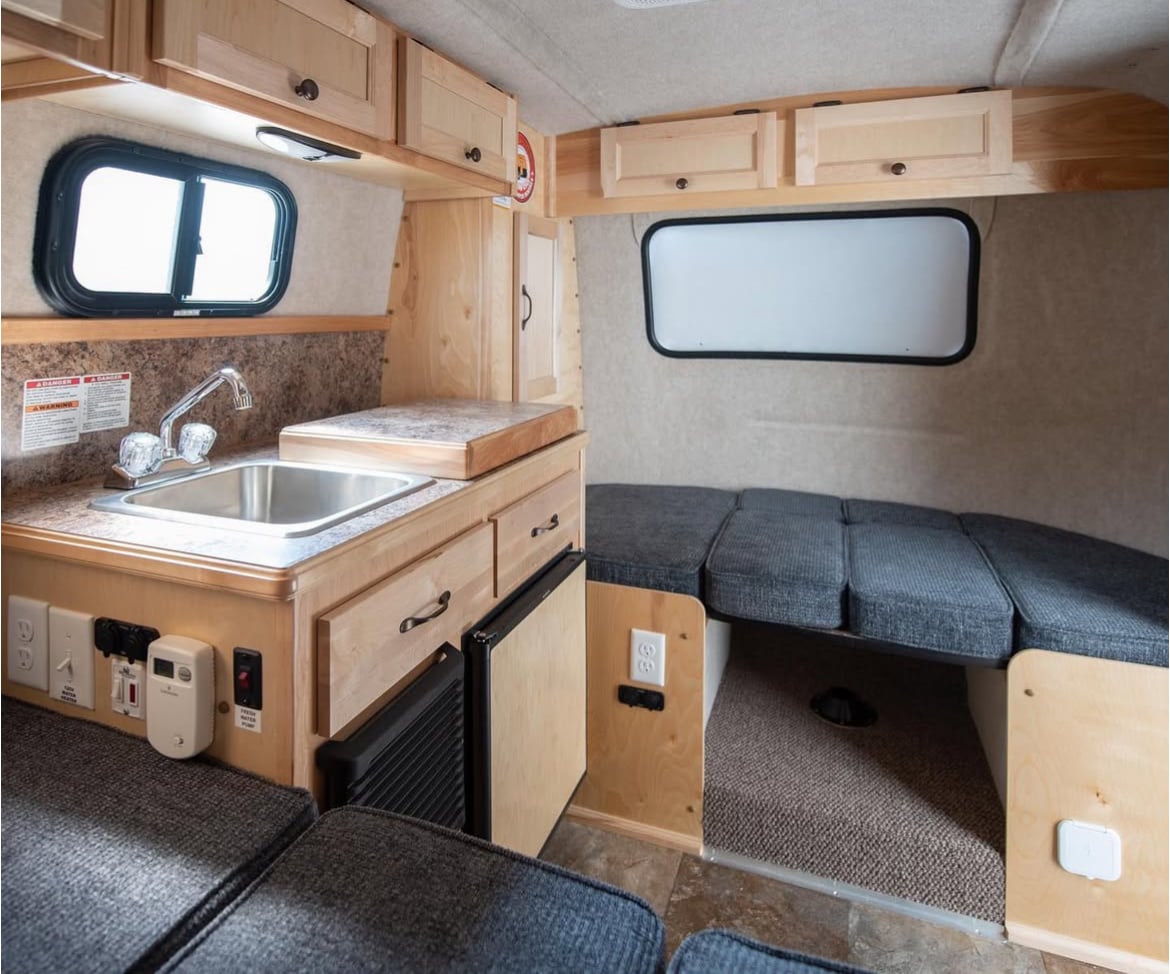 An interior view of a camper showcases wooden cabinetry and a compact kitchen area. A stainless steel sink is positioned next to a counter space. Folded seating creates an adaptable space, with a dark carpet covering the floor and a window allowing natural light to enter.