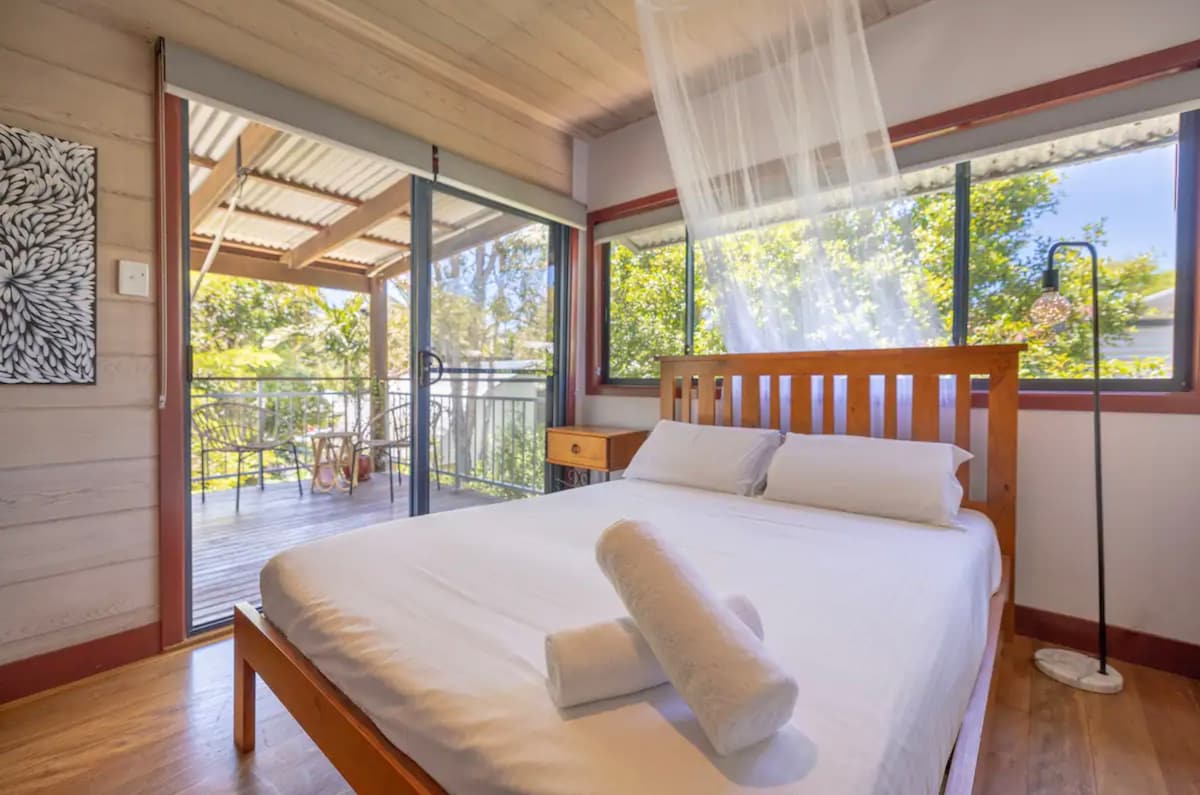 A serene bedroom features a spacious bed with freshly laundered linens and rolled towels. Sliding glass doors open to a private balcony, providing natural light and views of the surrounding trees. A minimalist floor lamp stands beside the bed, complementing the tranquil ambiance.
