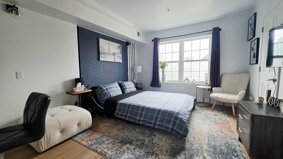 A spacious bedroom features a large window with natural light illuminating the room. A neatly made bed with a checked blanket is positioned centrally, accompanied by a comfortable armchair and a side table. Decorative elements include a blue accent wall and a patterned area rug.